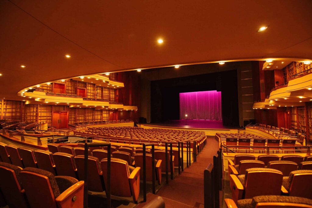 View of the Sandler theater with rows of seats, a wooden interior, and a stage with a pink curtain lit from above.