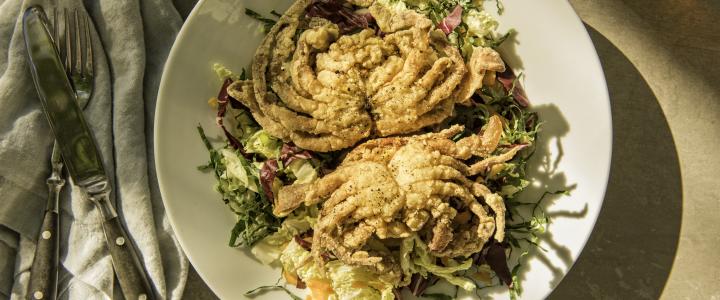 Two fried soft-shell crabs on a bed of mixed greens, served on a white plate with a fork and knife on a gray napkin beside it.