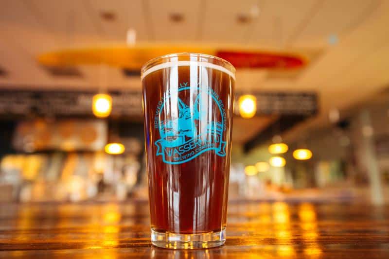 A pint glass of beer on a wooden table in a bar, with a logo on the glass. Blurred background with warm lighting.