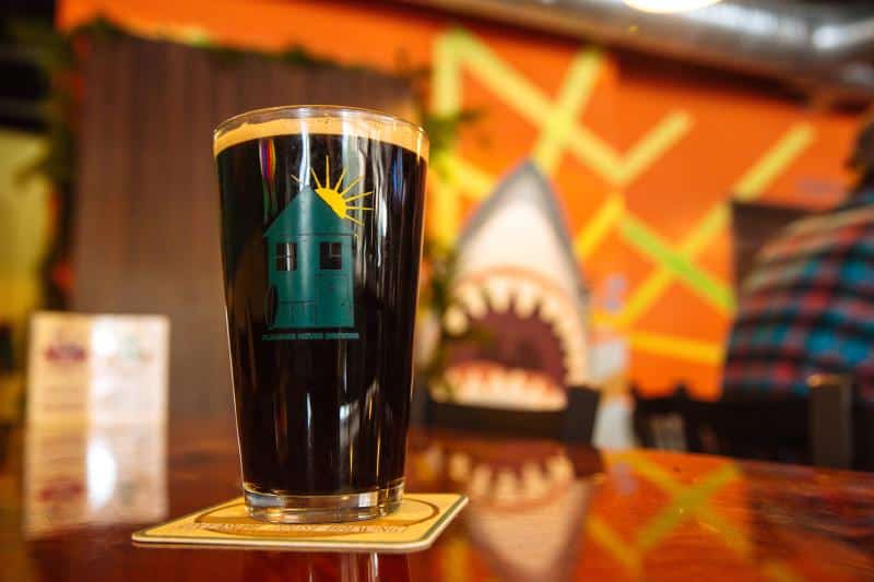 A glass of dark beer with a house logo sits on a coaster atop a wooden table. In the background, there's a colorful mural featuring a shark with an open mouth.