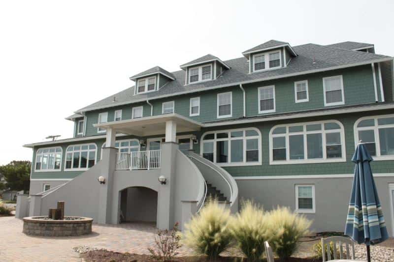 A large green and gray building with arched windows, an elevated entrance with stairs, and a circular area with a fire pit in the foreground. A closed umbrella stands nearby.