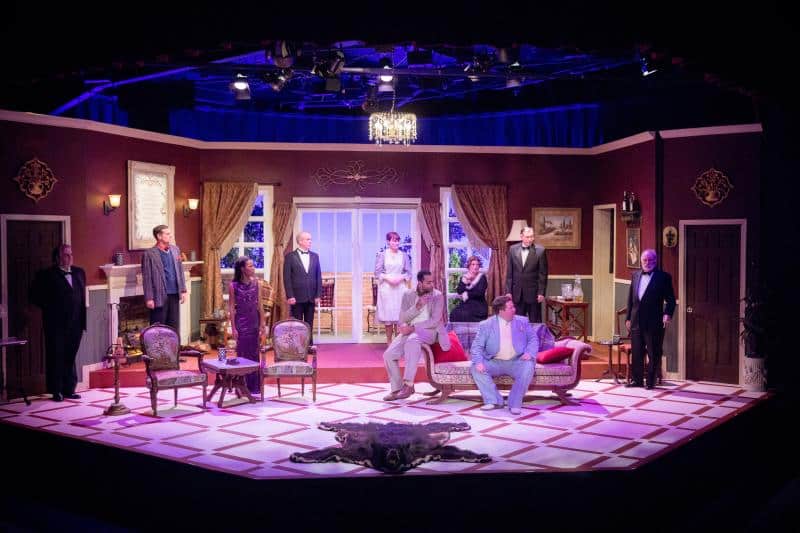 A stage play set in a living room with nine actors in various positions, featuring furniture, curtains, and a chandelier above, under stage lighting.