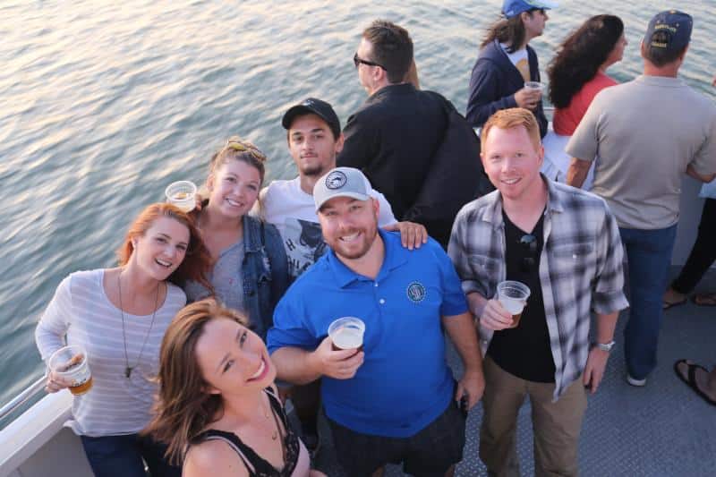 A group of people smiling and holding drinks on a boat with water in the background.