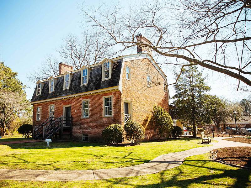 A historic brick house with a gambrel roof, surrounded by trees and a grassy lawn under a clear blue sky.