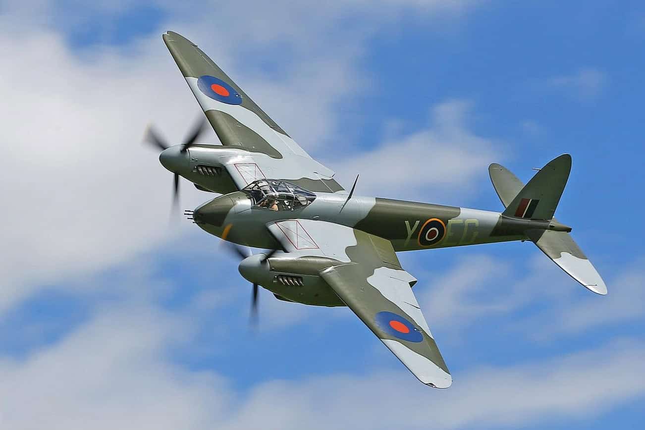World War II-era fighter plane in flight, displaying RAF roundels on wings against a blue sky with clouds.