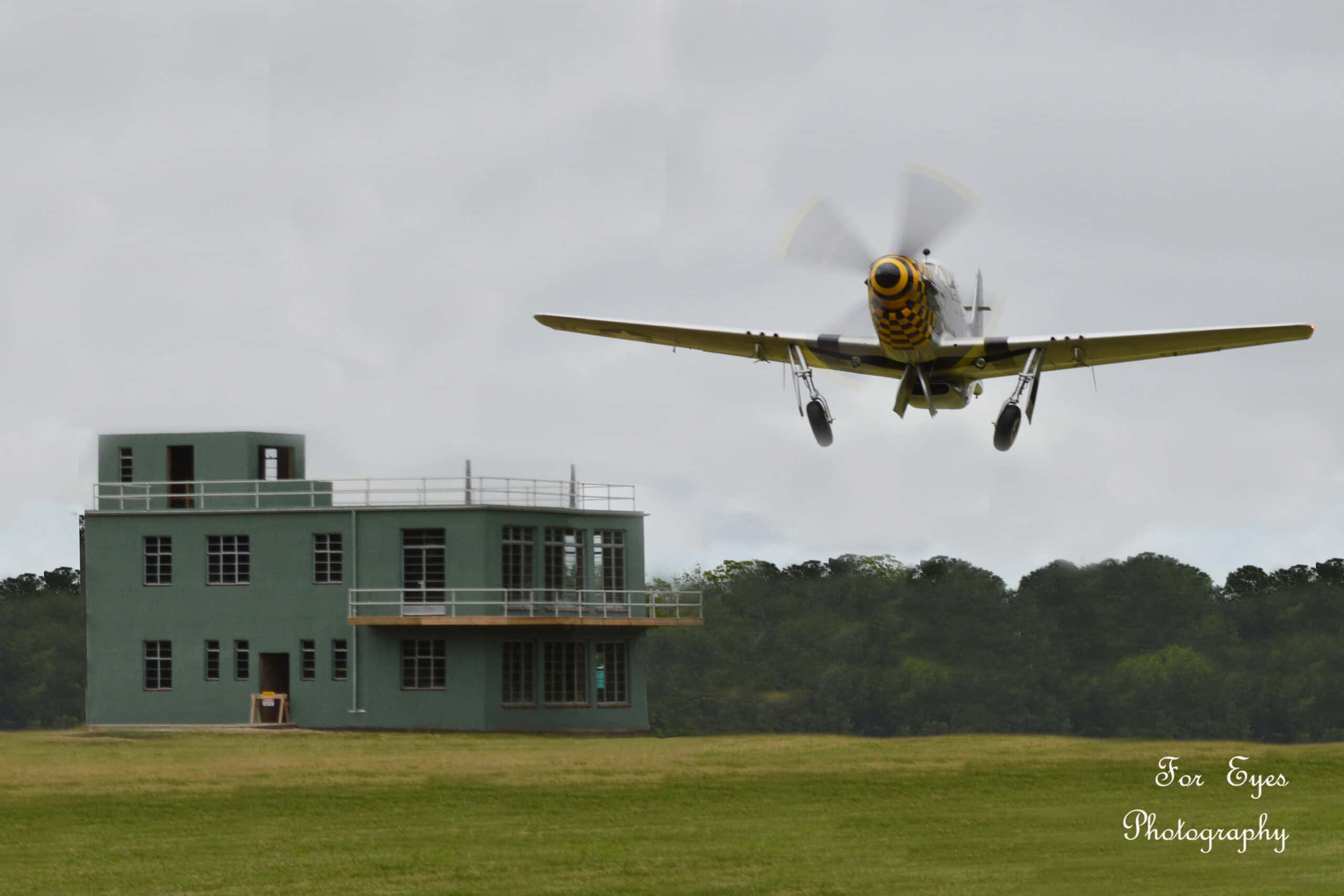 A vintage aircraft flies low near a two-story green building, surrounded by grass and trees under a cloudy sky.