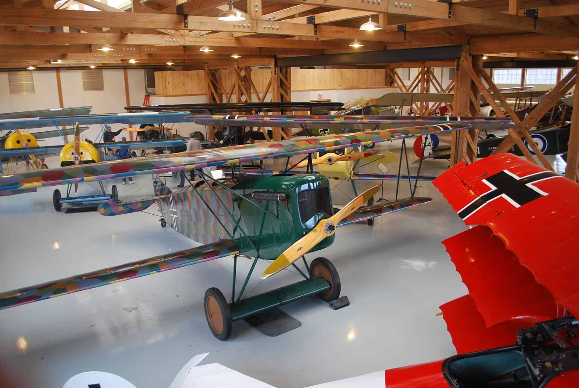A collection of vintage biplanes displayed in a wooden hangar, including a green plane with multicolored wings and a red plane with emblematic markings.