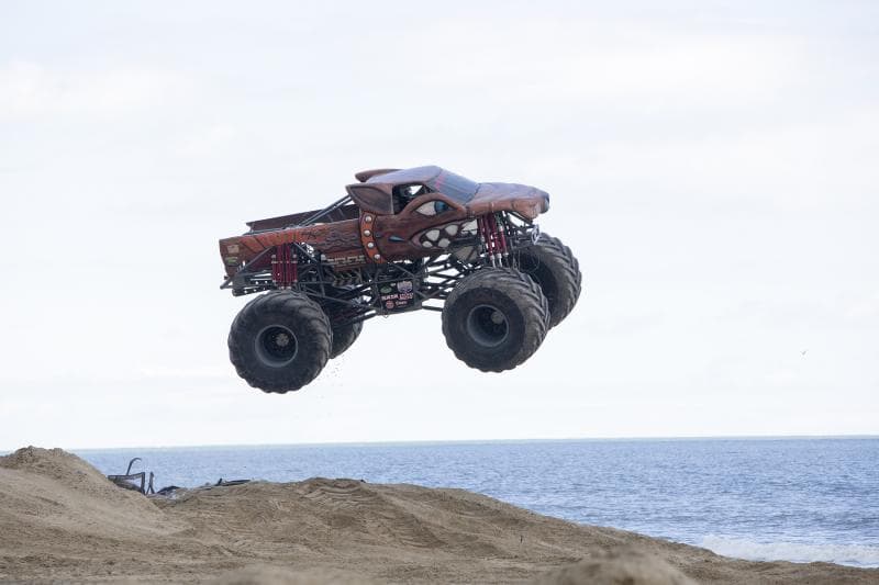 A monster truck with a brown, aerodynamic body jumps over a sandy ramp near the Virginia Beach oceanfront.