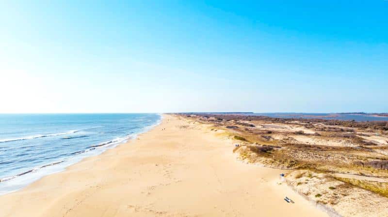 Aerial view of a sandy beach stretching alongside the ocean, with sparse vegetation on the dunes under a clear blue sky.