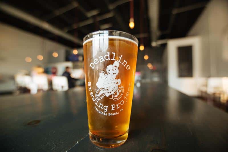 A pint glass with a gold-colored beer, labeled "Deadline Brewing Project." It rests on a table in a bar with blurred lights in the background.