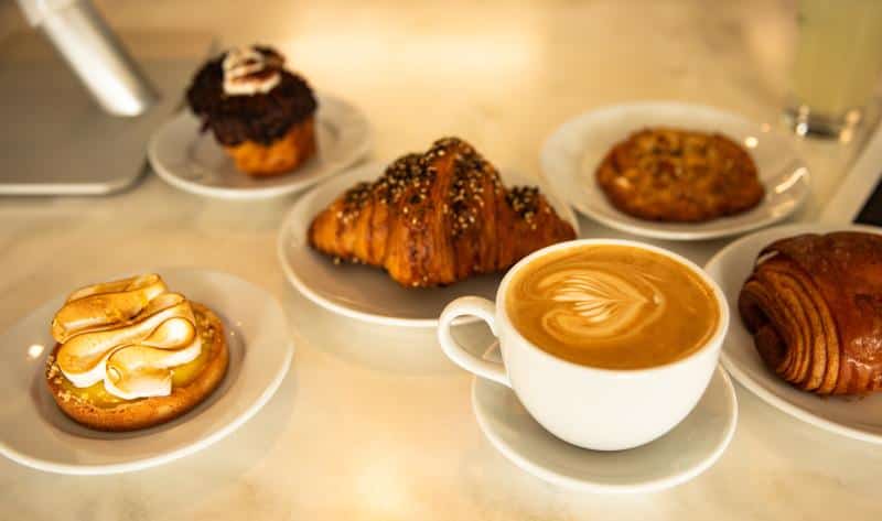 A cappuccino with latte art is surrounded by a lemon tart, a chocolate cupcake, a sesame croissant, a cookie, and a chocolate croissant on white plates.