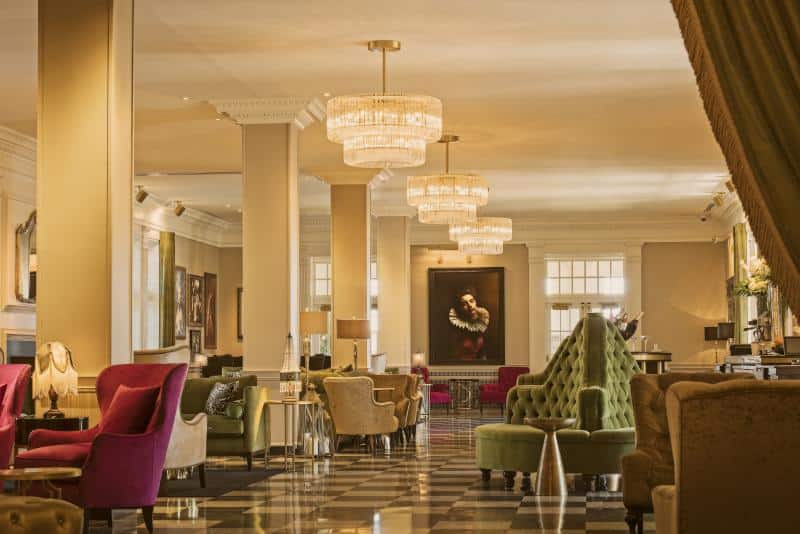 Elegant hotel lobby with plush chairs, chandeliers, and a checkered floor. Artwork and framed pictures adorn the walls, creating a sophisticated atmosphere.