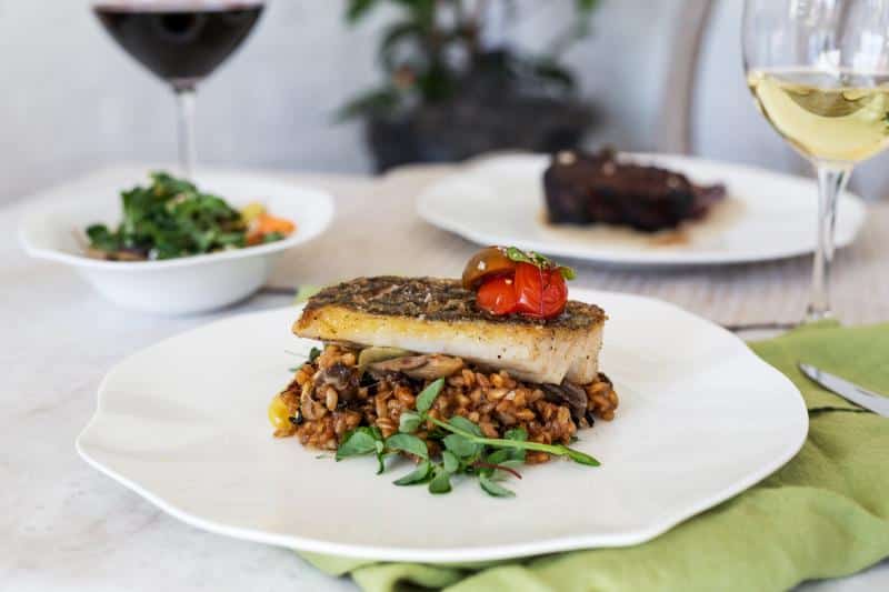 A plated dish featuring grilled fish on mushroom risotto, garnished with a cherry tomato and herbs. Wine glasses and another dish are visible in the background.