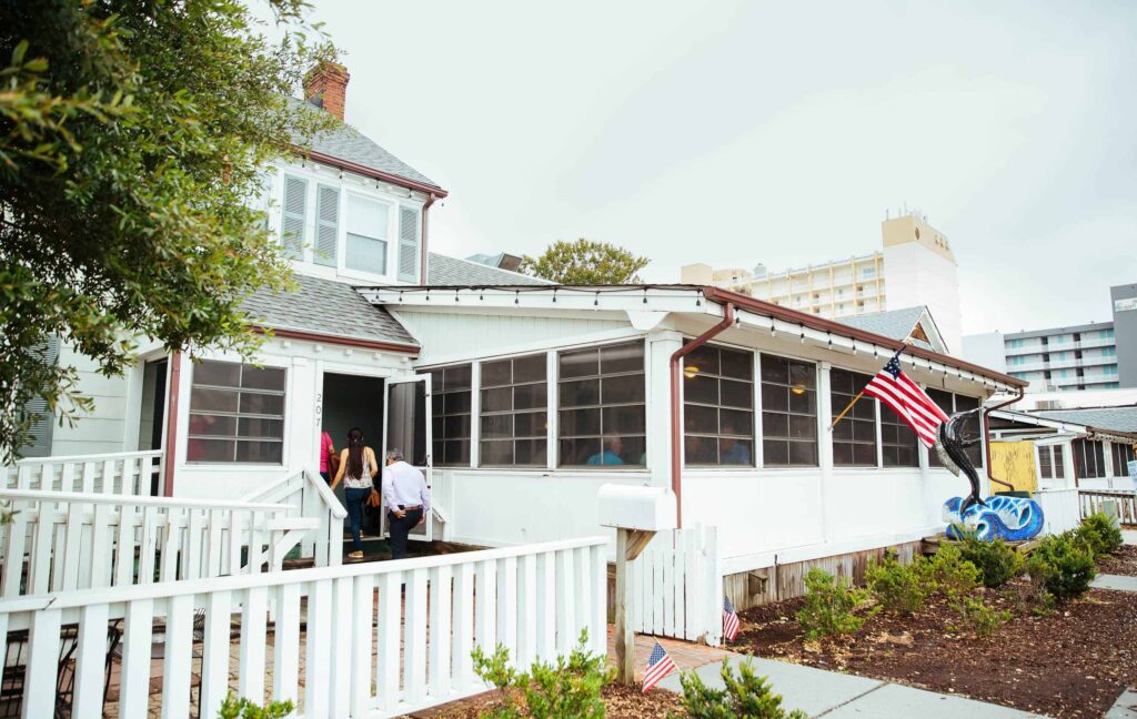 People enter Doc Taylor’s, a white coastal cottage-style restaurant near the Virginia Beach Oceanfront, with an American flag displayed outside.