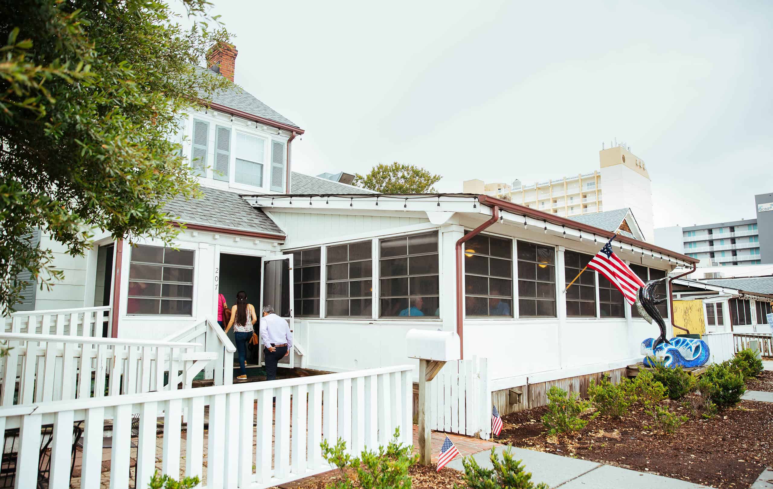 People entering a white, single-story house with a porch and an American flag.