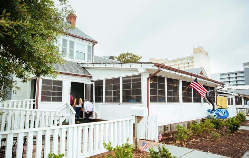 Two people enter a white house with a porch and an American flag. The building is surrounded by greenery and other structures are visible in the background.