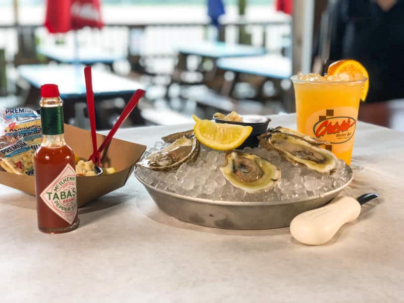 A plate of raw oysters on ice with lemon, a drink with an orange slice, hot sauce, crackers, and a cocktail sauce in a cup on a table.