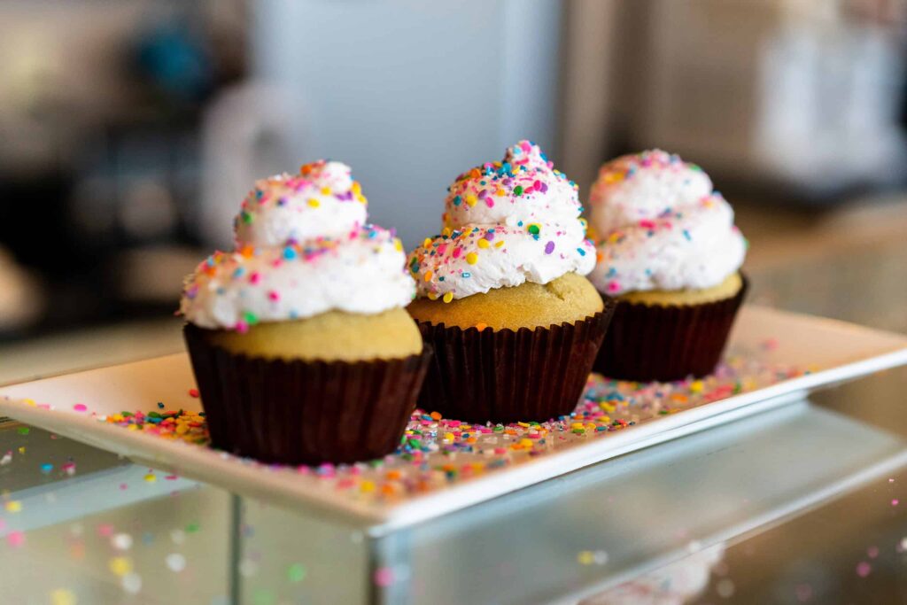 Three vanilla cupcakes with white frosting and colorful sprinkles on a rectangular white plate.