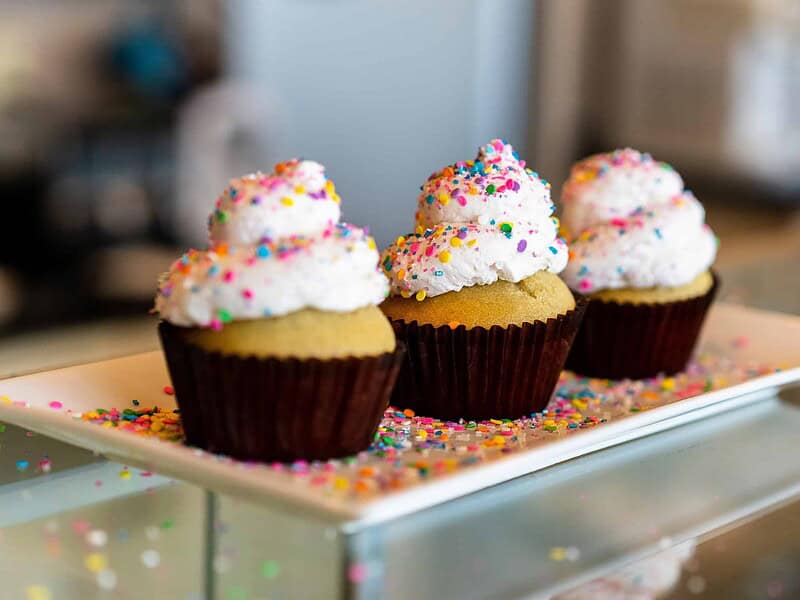Three vanilla cupcakes with white frosting and colorful sprinkles on a rectangular white plate.