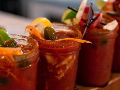 Four glasses of tomato-based cocktails garnished with lime wedges, carrots, olives, and spices sit on a wooden tray.