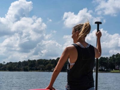 A woman with a ponytail stands on a paddleboard holding a paddle, looking out at a lake with trees in the background under a partly cloudy sky.