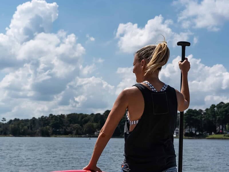 A woman with a ponytail stands on a paddleboard holding a paddle, looking out at a lake with trees in the background under a partly cloudy sky.