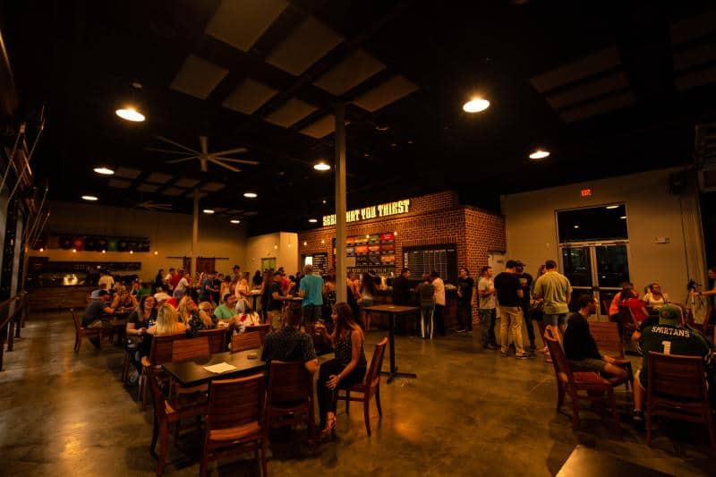 A dimly lit bar interior with patrons seated at tables and standing in line at the counter. Wooden chairs and tables are scattered around the room.