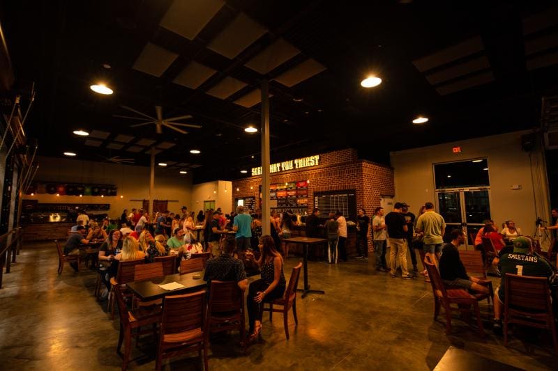 A busy indoor brewery taproom with patrons seated at wooden tables and in line at the bar. The space has a high ceiling and dim lighting, creating a warm atmosphere.