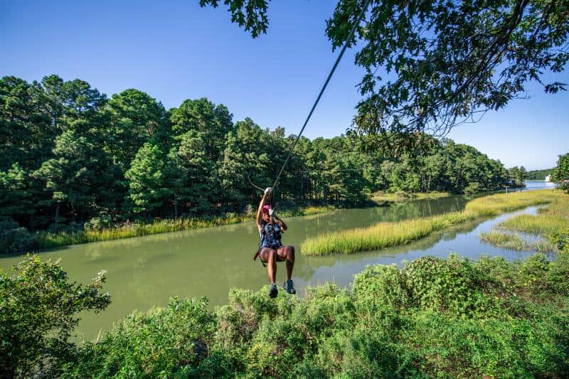 Person ziplining over a green, scenic river surrounded by lush trees under a clear blue sky.