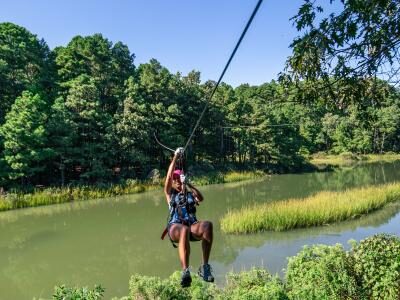 Person zip-lining over a body of water surrounded by lush green trees and vegetation under a clear blue sky.
