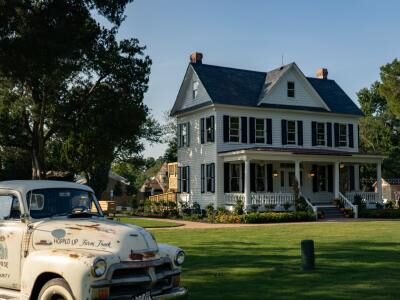 A vintage white truck is parked on a lush green lawn in front of a large white two-story house with a porch, surrounded by trees under a clear blue sky.