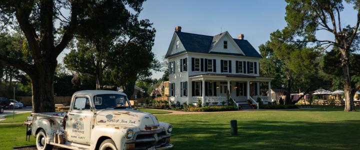 A vintage white truck is parked on a lush green lawn in front of a large white two-story house with a porch, surrounded by trees under a clear blue sky.