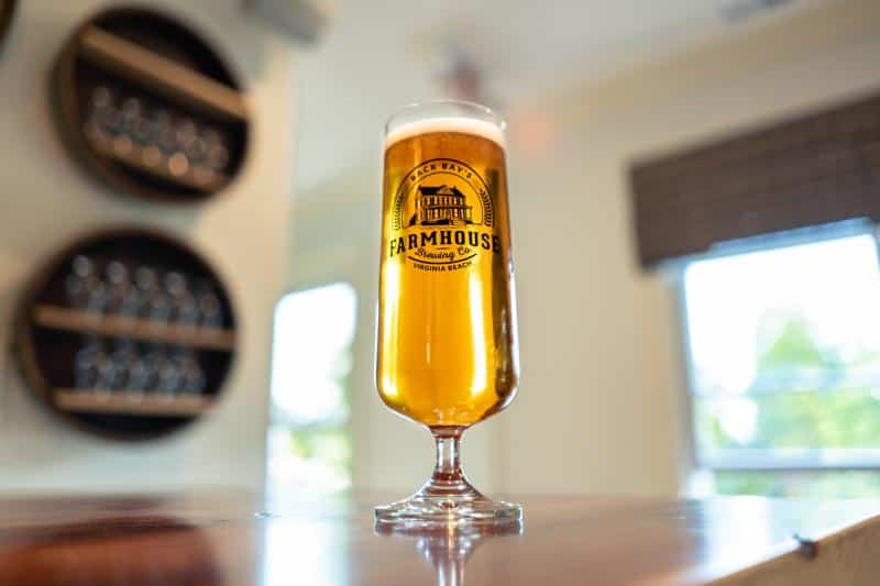 A tall glass filled with beer sits on a wooden counter. The glass has "Farmhouse Brewing Co." printed on it. In the background, there are windows and round shelves with glasses.