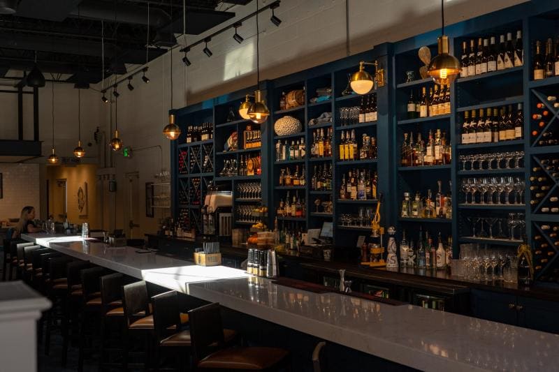 Bar interior with a long white countertop, dark blue shelves filled with bottles and glasses, and warm hanging lights illuminating the space.