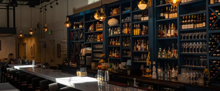 A dimly lit bar with a long counter, barstools, and shelves stocked with various bottles and glasses. Warm pendant lights hang from the ceiling.
