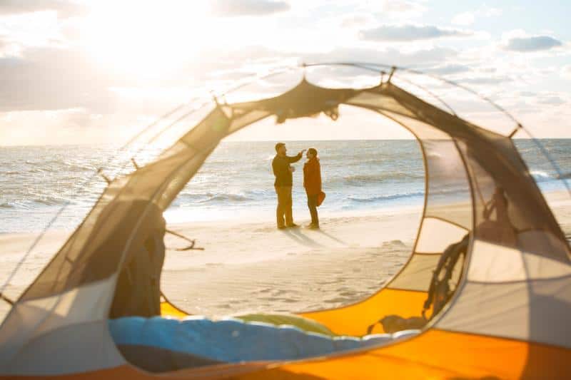 View through a tent opening of two people standing and interacting on a sandy beach with the ocean and a bright sun in the background.