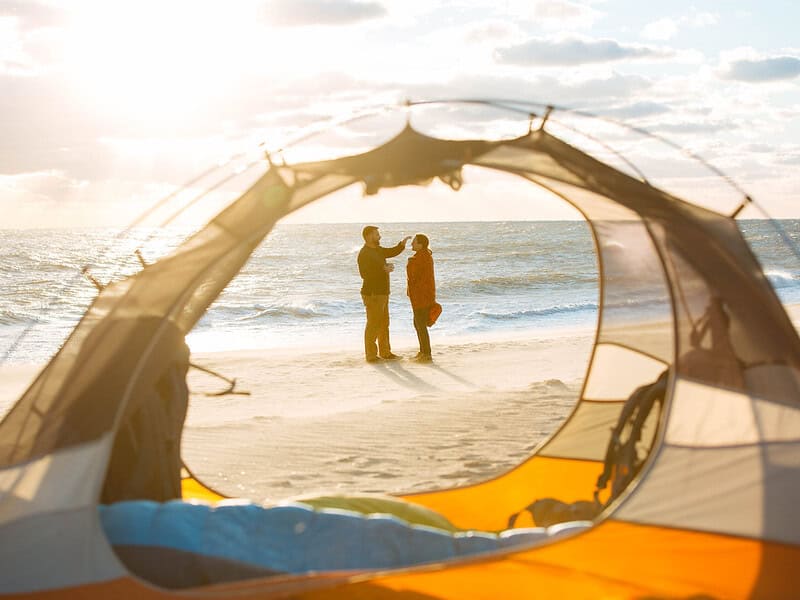 A couple stands on a sunlit beach near the ocean, seen through the opening of a tent.