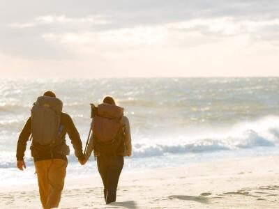 Two people with backpacks walk hand in hand along a sandy beach, with the ocean in the background under a cloudy sky.