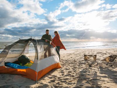 Two people stand near a tent on a sandy beach. The ocean is in the background under a cloudy sky. Two chairs and a small table are set up nearby.