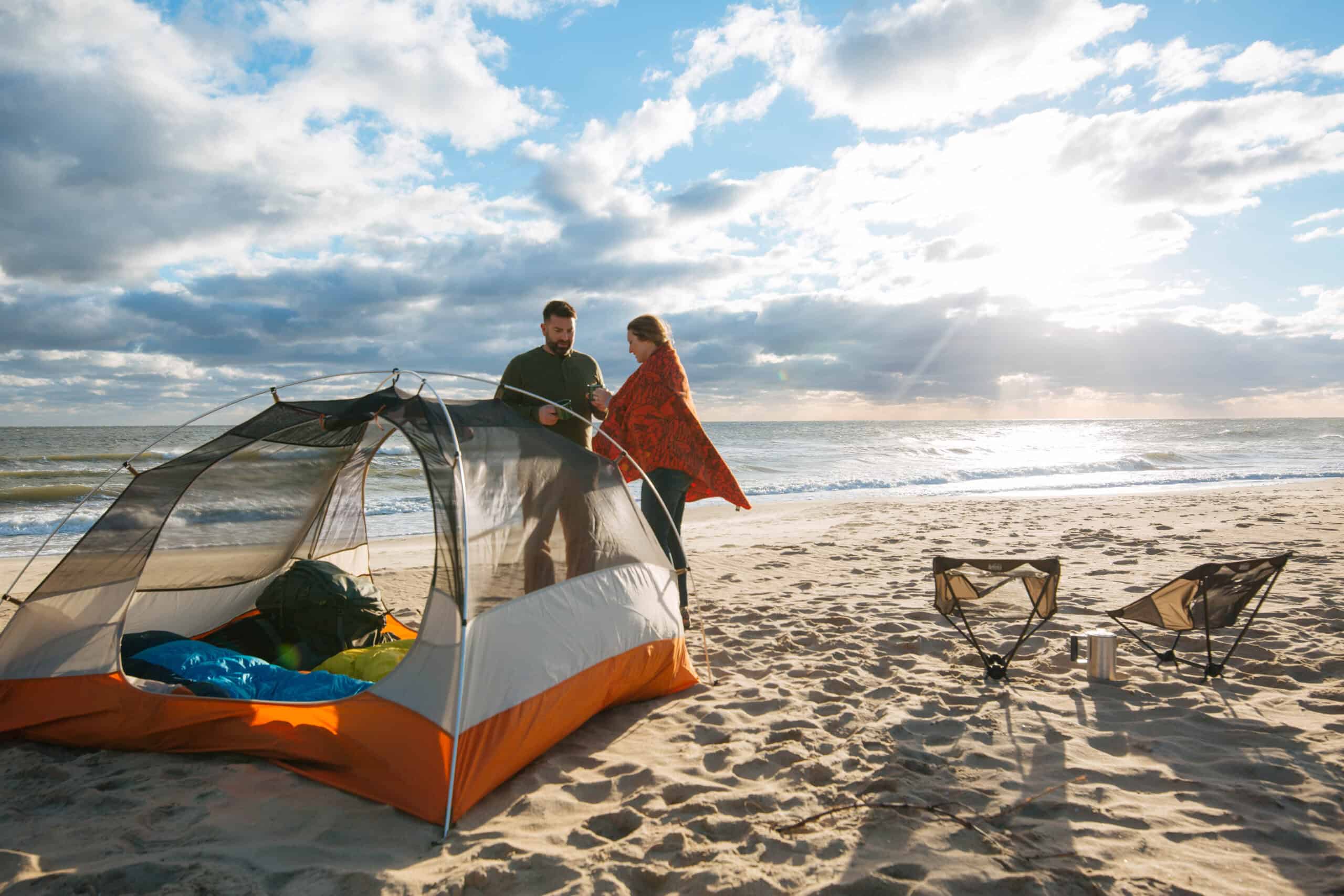 Two people stand near a tent on a sandy beach. The ocean is in the background under a cloudy sky. Two chairs and a small table are set up nearby.