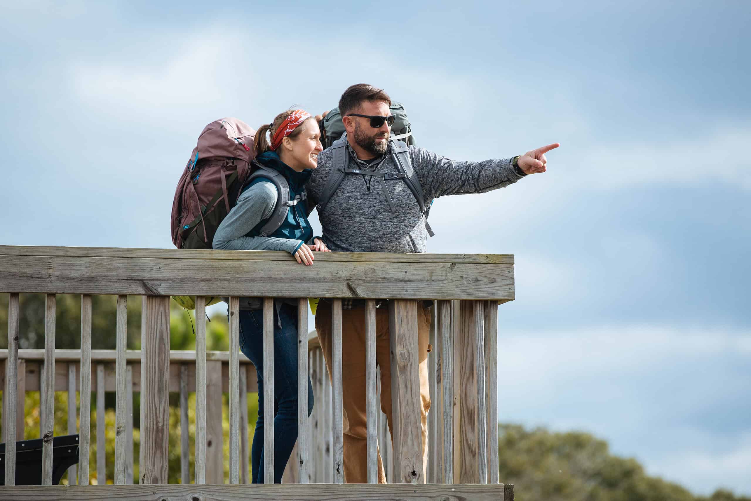 
Two people with backpacks stand on a wooden deck, with one pointing into the distance. They are outdoors under a cloudy sky.