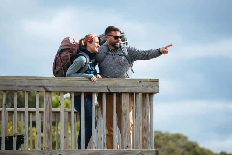 Two people with backpacks stand on a wooden deck. The man points into the distance, while the woman looks in the same direction. They appear to be outdoors with a cloudy sky background.