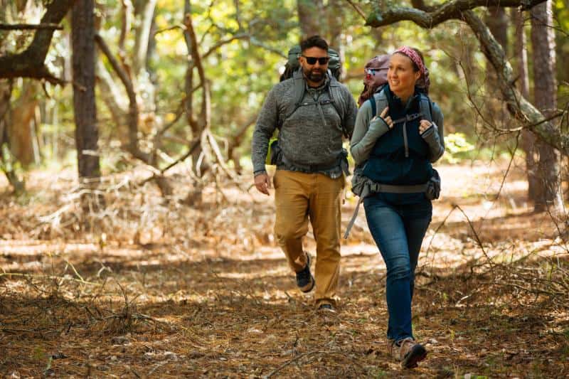 Two people hiking in a forest, carrying backpacks. The woman in front is smiling, while the man follows behind wearing sunglasses. Trees and fallen branches are visible in the background.