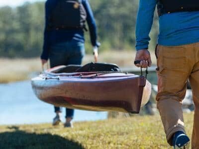 Two people carry a kayak towards a body of water, surrounded by trees and grass.