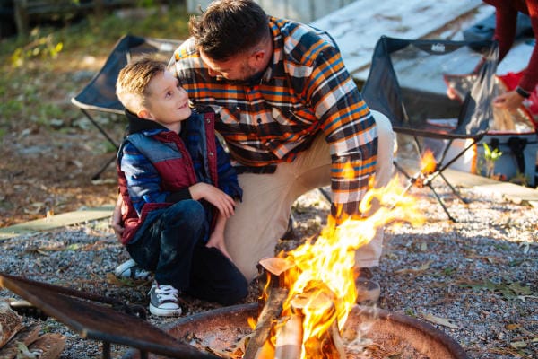 Man and child sitting by a campfire, with camping chairs in the background.
