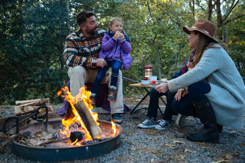 A family sitting around a campfire. A man and a woman are roasting marshmallows, while a child in a purple jacket eats one. The scene is set in a wooded area, with camping gear nearby.