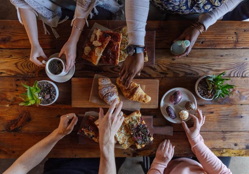 People sharing pastries, coffee, and small dishes at a wooden table. Some hands reach for food while others hold beverages.