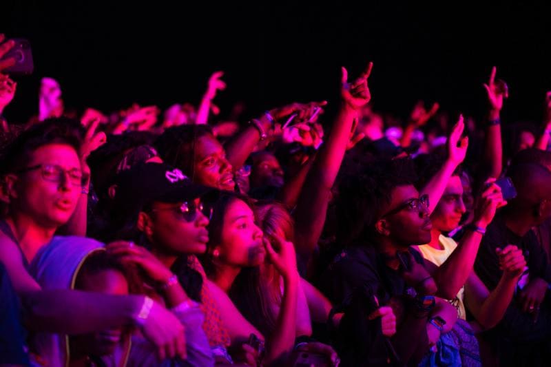A crowd of people watches a concert at night, with some holding up their phones. The scene is illuminated by pink and purple lighting.