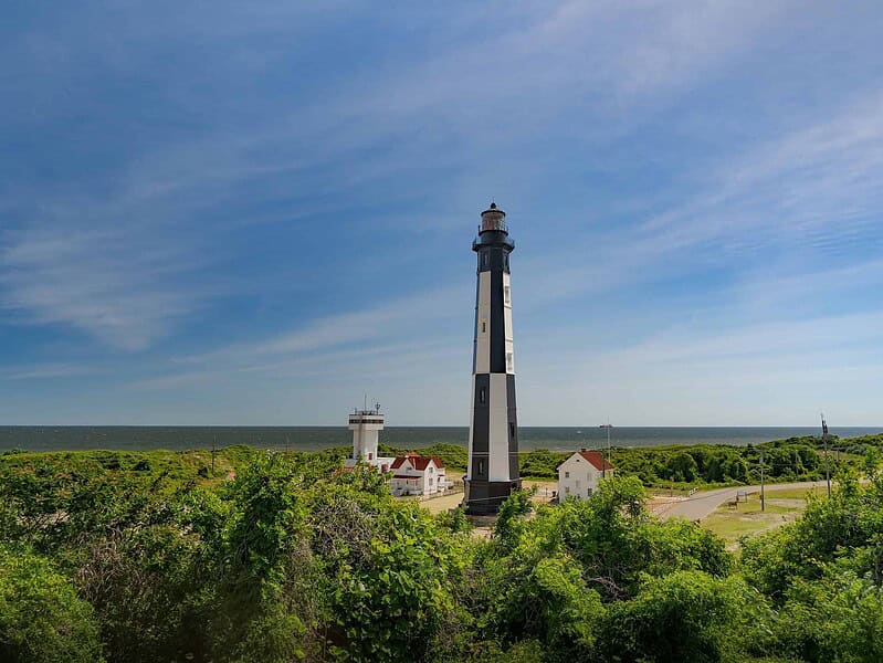 Lighthouse stands tall amidst green foliage with smaller buildings nearby, under a blue sky overlooking the ocean in the background.