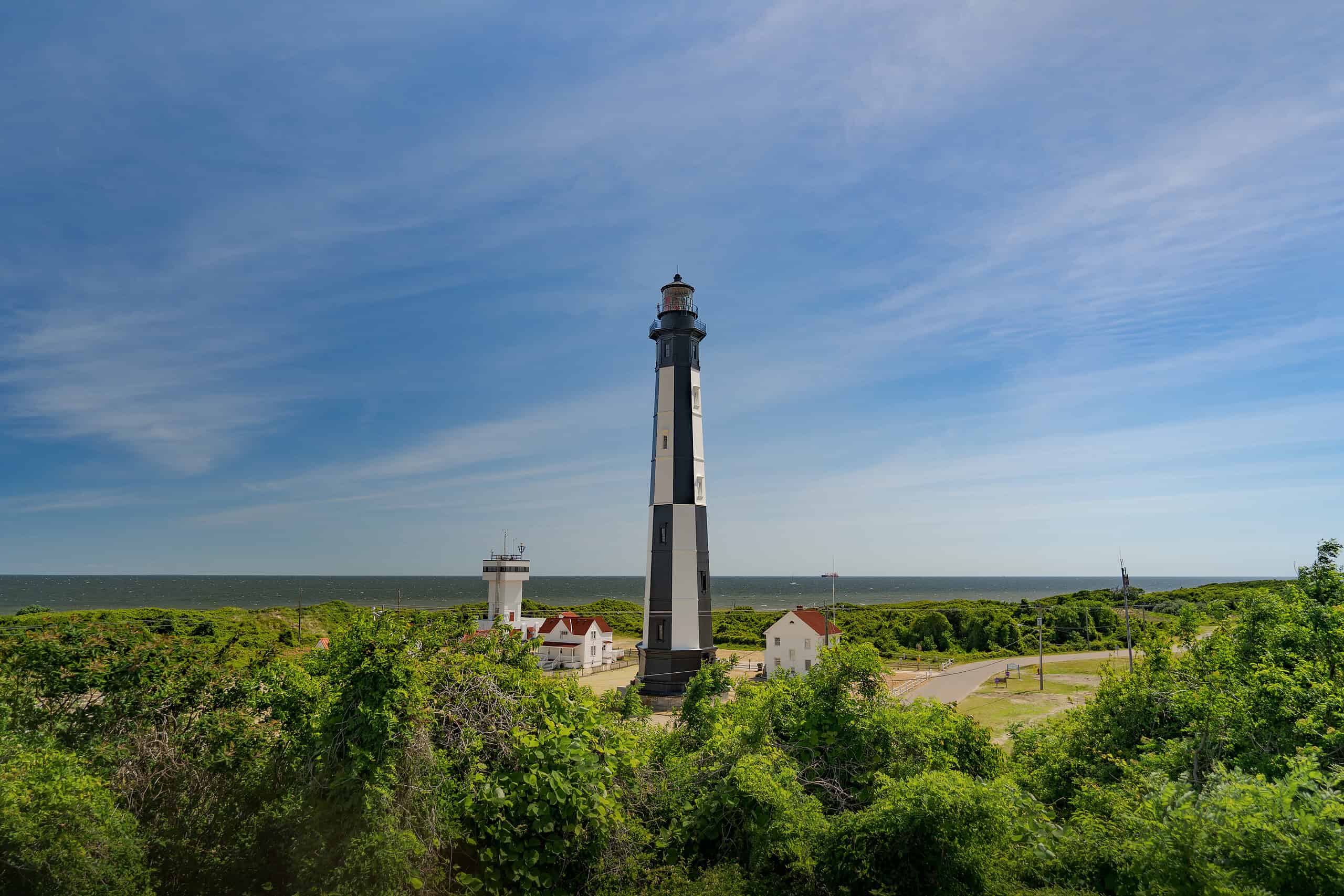 Lighthouse stands tall amidst green foliage with smaller buildings nearby, under a blue sky overlooking the ocean in the background.